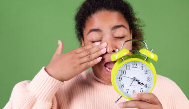 Tired woman yawning and holding green alarm clock representing disrupted circadian rhythm and morning fatigue.