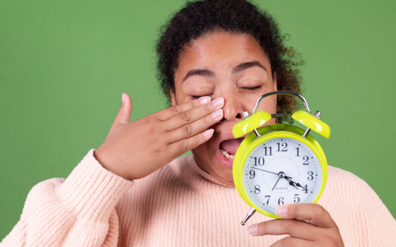 Tired woman yawning and holding green alarm clock representing disrupted circadian rhythm/ weight loss and morning fatigue.
