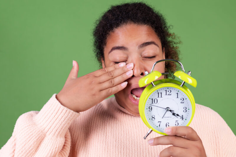 Tired woman yawning and holding green alarm clock representing disrupted circadian rhythm and morning fatigue.