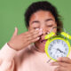 Tired woman yawning and holding green alarm clock representing disrupted circadian rhythm and morning fatigue.